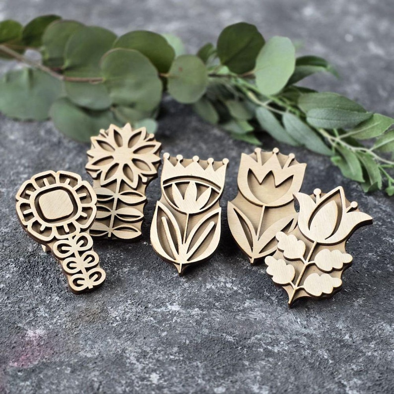 May include: Five wooden floral stamps in various designs, including a sunflower, tulip, and other stylized flowers. The stamps are light brown and arranged on a grey surface with green foliage in the background. These stamps are ideal for crafting and DIY projects.