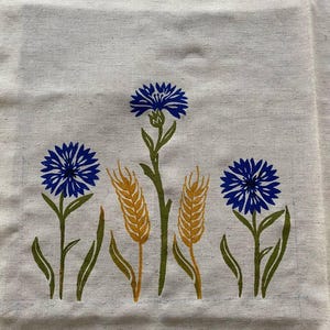 May include: A cream-coloured fabric featuring a floral design with blue cornflowers and golden wheat stalks. The flowers have detailed petals and are set against green stems and leaves. The design is printed on a textured, natural-looking material.