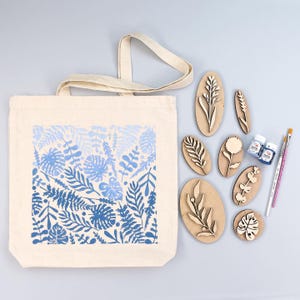 May include: A cream-colored tote bag with a blue and white floral design. Wooden stamps with various botanical shapes, two small paint bottles, and paint brushes are arranged next to the bag. The design includes leaves and ferns.