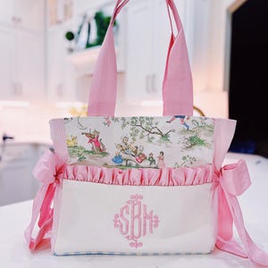 K&ouml;nnte beinhalten: Eine rosa und wei&szlig;e Tragetasche mit Blumendruck und rosa R&uuml;schenbesatz. Die Tasche hat eine wei&szlig;e Tasche mit einem rosa Monogramm, das "SBM" sagt.