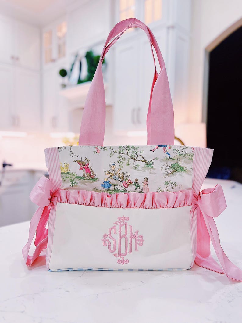 K&ouml;nnte beinhalten: Eine rosa und wei&szlig;e Tragetasche mit Blumendruck und rosa R&uuml;schenbesatz. Die Tasche hat eine wei&szlig;e Tasche mit einem rosa Monogramm, das "SBM" sagt.