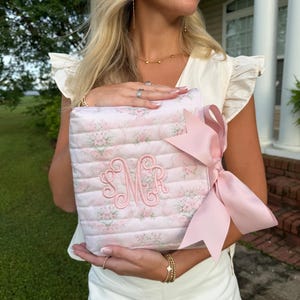 May include: A square, quilted fabric pouch featuring a light pink floral pattern and horizontal stitching. The center displays an embroidered monogram with the letters SMR in pink thread. A large, solid light pink satin ribbon bow is attached.
