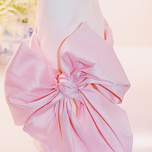 K&ouml;nnte beinhalten: Eine wei&szlig;e Einkaufstasche mit einer gro&szlig;en rosa Schleife aus Satin. Die Schleife ist in einem Knoten gebunden und die Enden der Schleife schweben.