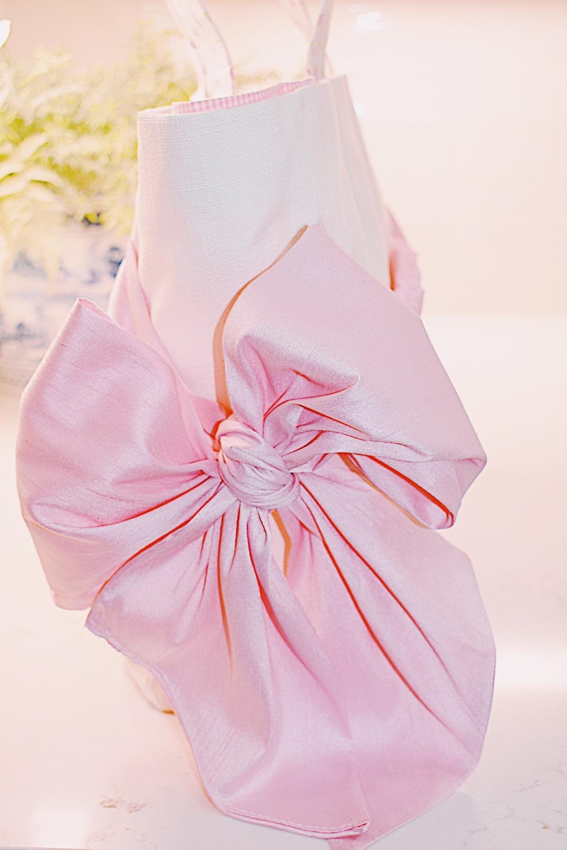 K&ouml;nnte beinhalten: Eine wei&szlig;e Einkaufstasche mit einer gro&szlig;en rosa Schleife aus Satin. Die Schleife ist in einem Knoten gebunden und die Enden der Schleife schweben.