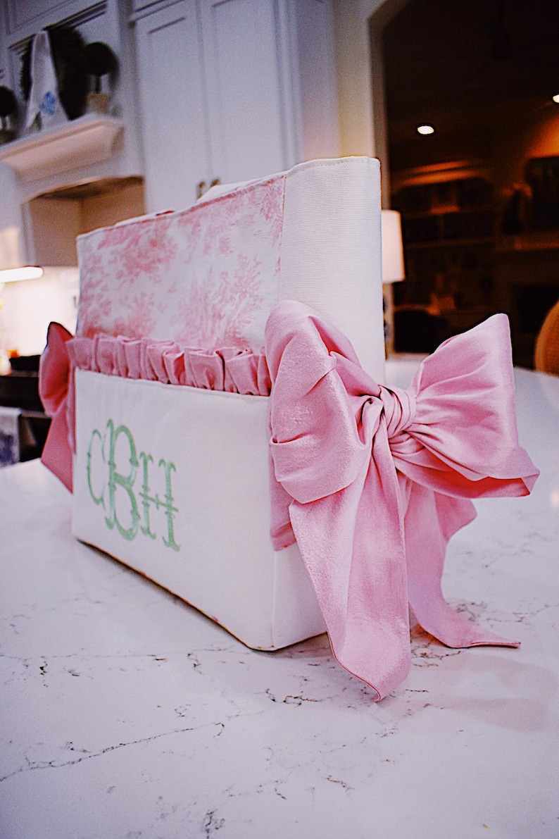 K&ouml;nnte beinhalten: Ein wei&szlig;er und rosa Aufbewahrungskorb mit einem gr&uuml;nen Monogramm "CBH" und einer gro&szlig;en rosa Schleife aus Satin. Der Korb hat einen gekr&auml;uselten Rand und ein Blumenmuster.