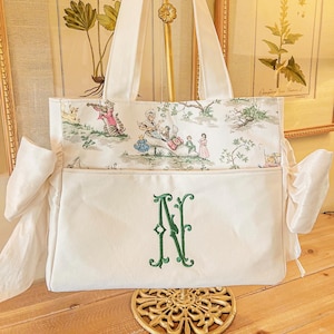 K&ouml;nnte beinhalten: Eine wei&szlig;e Canvas-Tragetasche mit einem gr&uuml;nen gestickten Monogramm "N" auf der Vorderseite. Die Tasche hat ein Futter mit Blumendruck und zwei wei&szlig;e Stoffschleifen an den Seiten.