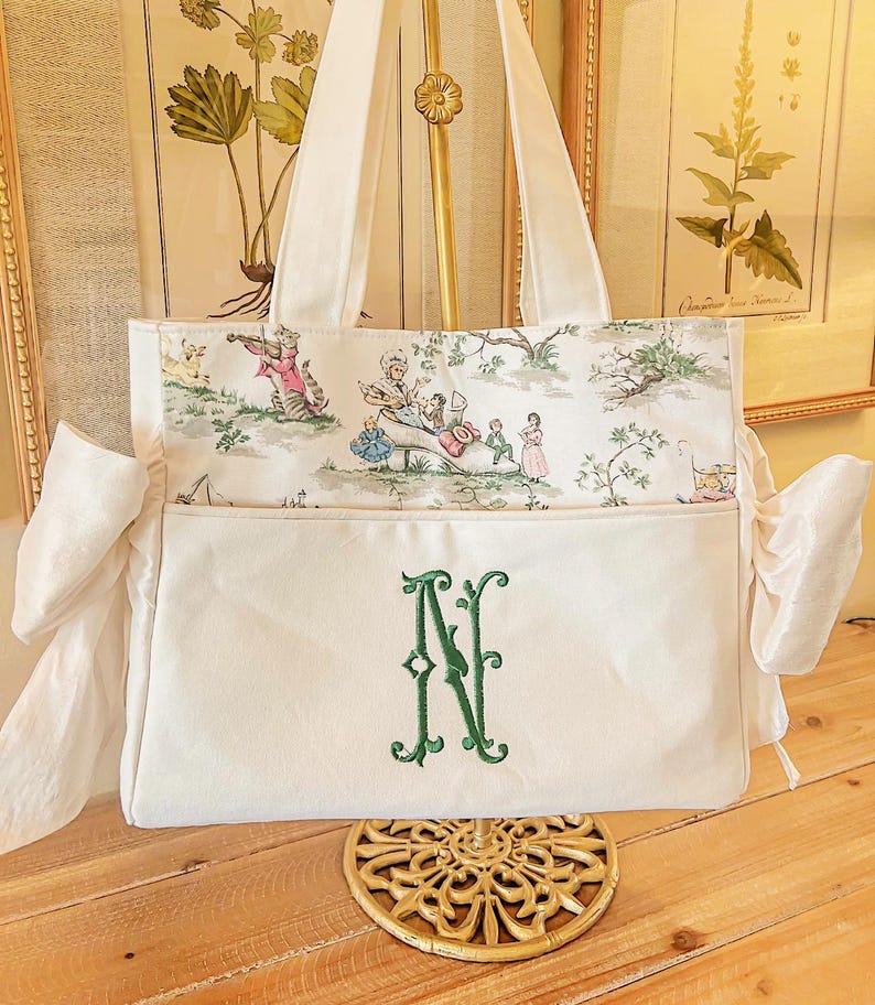 K&ouml;nnte beinhalten: Eine wei&szlig;e Canvas-Tragetasche mit einem gr&uuml;nen gestickten Monogramm "N" auf der Vorderseite. Die Tasche hat ein Futter mit Blumendruck und zwei wei&szlig;e Stoffschleifen an den Seiten.
