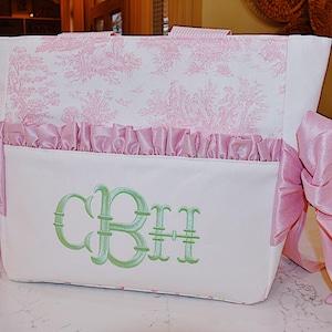 K&ouml;nnte beinhalten: Eine wei&szlig;e Tragetasche mit einem rosafarbenen Blumendruck und einem gr&uuml;nen Monogramm "CBH", das auf der Vorderseite gestickt ist. Die Tasche hat an jeder Seite rosafarbene Satinb&auml;nder.