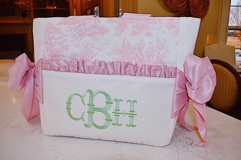 K&ouml;nnte beinhalten: Eine wei&szlig;e Tragetasche mit einem rosafarbenen Blumendruck und einem gr&uuml;nen Monogramm "CBH", das auf der Vorderseite gestickt ist. Die Tasche hat an jeder Seite rosafarbene Satinb&auml;nder.