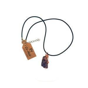 May include: A handcrafted necklace with a dark blue stone encased in copper wire, hanging from a black cord. The necklace includes a small tag with the text "Co-Heir Creations". The stone has a natural, uneven texture.