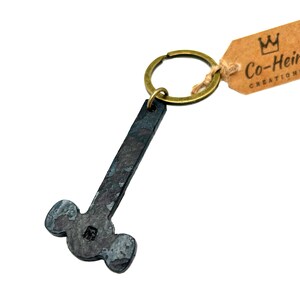 May include: A black metal hammer keychain with a brass key ring. The keychain is attached to a brown tag with the text "Co-Heir Creations" and a crown logo.