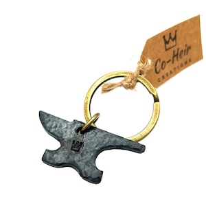 May include: A black metal anvil keychain with a brass key ring. The anvil has a bottle opener on the side and a crown logo on the front. The keychain is attached to a brown paper tag with the text "Co-Heir Creations" and a crown logo.