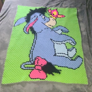 May include: A handmade, crocheted blanket featuring Eeyore from Winnie the Pooh. The blanket showcases Eeyore in shades of blue, pink, and black, with a pink bow and a butterfly. The background is bright green.