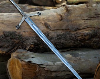May include: A silver and steel sword with a cross guard and a decorative handle. The sword is lying on a piece of wood.