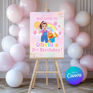 Op de afbeelding: Een feestbord op een houten ezel met de tekst "Welkom bij Olivia's 2e verjaardag!" met stripfiguren, een regenboog en ballonnen. Roze en witte ballonnen omringen het bord.