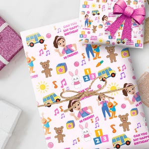 May include: Birthday wrapping paper in pink, featuring cartoon characters, musical notes, and the text "Can You Say Happy Birthday?" A gift is wrapped with twine and a pink bow.