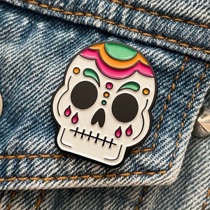 A skull enamel pin pinned on a denim jacket. The skull is white with green, gold, and pink accents on the top of the head and above the eyes. Pink tear drops fall from the eyes.