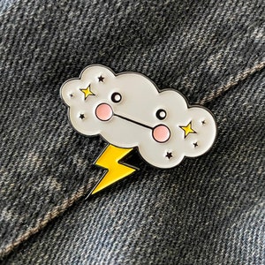 May include: A white enamel pin with a cartoon cloud with a lightning bolt underneath. The cloud has a smiling face with pink cheeks and black eyes. There are yellow stars around the cloud.