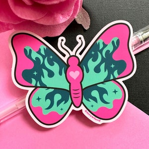 A die cut sticker of a pink and teal butterfly with a fire flame design in the wings. The butterfly's body is bright pink with a heart.