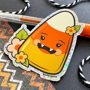 A die cut glitter sticker of a candy corn with a cute happy face and little fangs. There are peach and yellow flowers surrounding the candy corn.