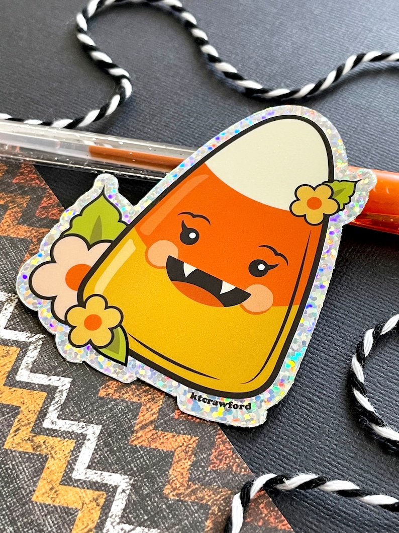 A die cut glitter sticker of a candy corn with a cute happy face and little fangs. There are peach and yellow flowers surrounding the candy corn.