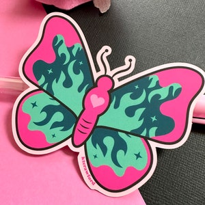 A die cut sticker of a pink and teal butterfly with a fire flame design in the wings. The butterfly's body is bright pink with a heart.