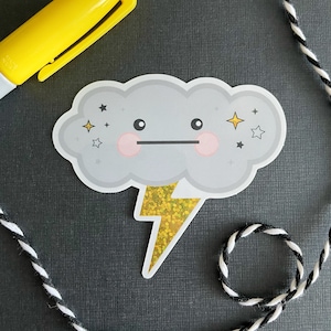 May include: A gray cloud with a cute cartoon face and a gold glitter lightning bolt. The cloud has two pink cheeks and black eyes. There are small gold stars around the cloud.