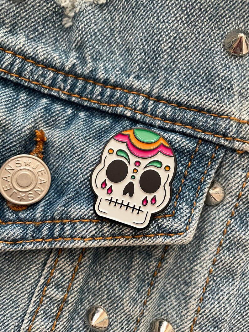 A skull enamel pin pinned on a denim jacket. The skull is white with green, gold, and pink accents on the top of the head and above the eyes. Pink tear drops fall from the eyes.