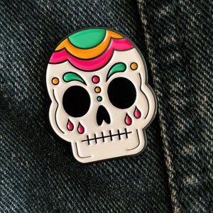 A skull enamel pin pinned on a denim jacket. The skull is white with green, gold, and pink accents on the top of the head and above the eyes. Pink tear drops fall from the eyes.