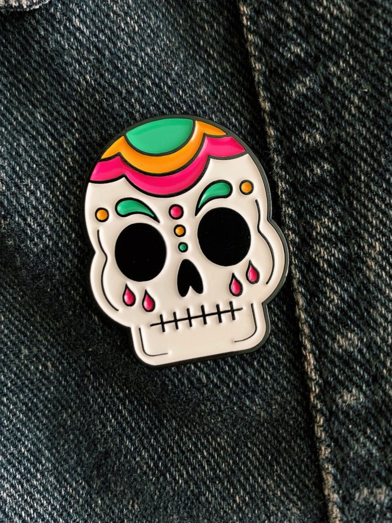 A skull enamel pin pinned on a denim jacket. The skull is white with green, gold, and pink accents on the top of the head and above the eyes. Pink tear drops fall from the eyes.