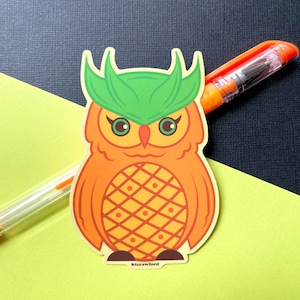 A die cut sticker of an orange owl that resembles a pineapple. The owl has yellow accents on its belly and around its green eyes. The top of its head is green.