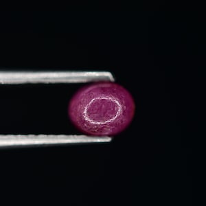 May include: A close-up of a deep pink gemstone held in tweezers against a black background.