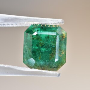 May include: A green emerald gemstone with inclusions, held by tweezers. The emerald is cut in a square shape with beveled edges.