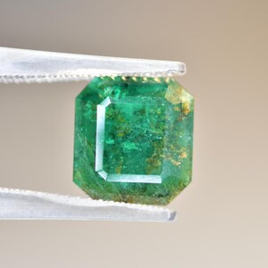 May include: A vibrant green emerald gemstone with inclusions, cut in a square emerald shape, held by tweezers.