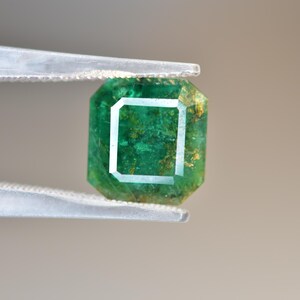 May include: A vibrant green emerald gemstone with inclusions, held in tweezers. The emerald has a square shape with cut facets.