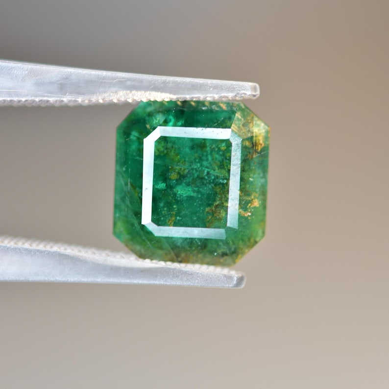 May include: A vibrant green emerald gemstone with inclusions, held in tweezers. The emerald has a square shape with cut facets.