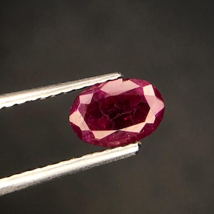 May include: A deep red oval-shaped gemstone with a faceted cut. The gemstone is held by a pair of tweezers.