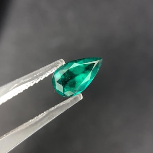 May include: A pear-shaped emerald gemstone with a deep green color. The gemstone is held in tweezers against a dark gray background.