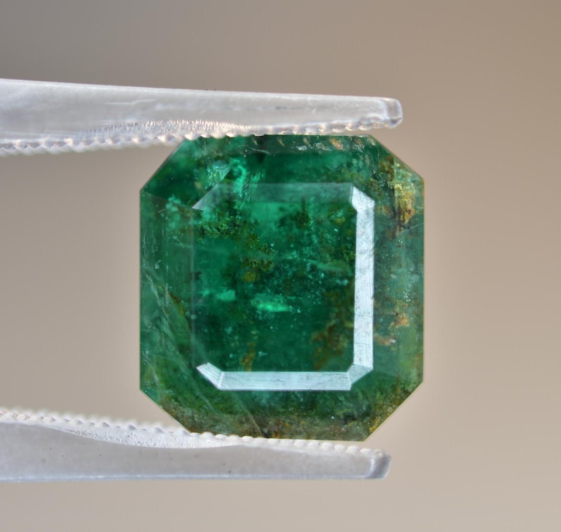 May include: A vibrant green emerald gemstone with a rectangular cut and visible inclusions. The emerald is held in tweezers.