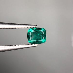 May include: A vibrant, cushion-cut emerald gemstone, held by a pair of silver tweezers. The emerald displays a deep green hue with internal inclusions, and is set against a neutral gray backdrop. The gemstone's facets reflect light, creating a sparkling effect.
