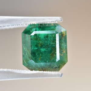 May include: A green emerald gemstone with inclusions, cut in an emerald shape, held in tweezers.