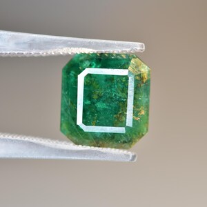 May include: A green emerald gemstone with inclusions, cut into a square shape with beveled edges. The emerald is held in tweezers.