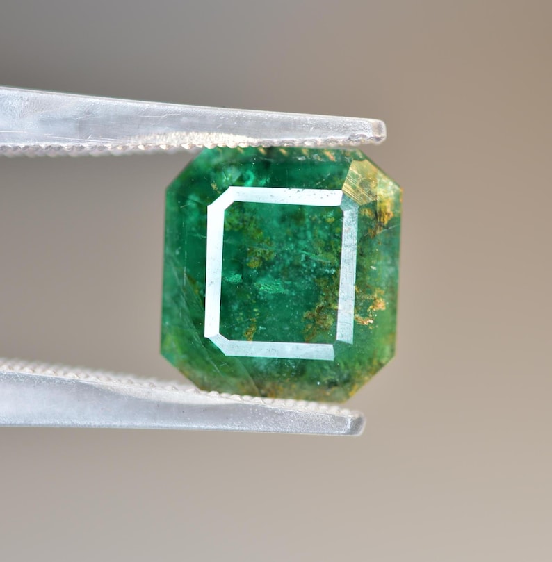 May include: A green emerald gemstone with inclusions, cut into a square shape with beveled edges. The emerald is held in tweezers.