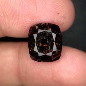 Pode incluir: Uma pedra preciosa castanha escura com um padrão de estrela cortado em sua superfície. A pedra preciosa é retangular com cantos arredondados. As facetas da estrela refletem a luz. A pedra tem uma aparência ligeiramente manchada.