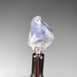 May include: A rough-cut, light blue gemstone is held by a silver metal tool. The gemstone has a crystalline structure with a slightly translucent appearance. The tool has a textured handle and a wire clasp holding the stone.