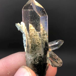 Pode incluir: Um cristal de quartzo transparente com inclusões verdes. O cristal é pontudo na parte superior e tem uma base áspera e natural.