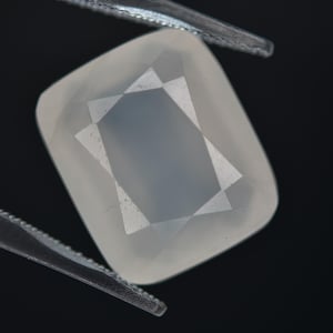 May include: A clear, faceted, white gemstone with a star-shaped pattern inside. The gemstone is held by tweezers against a black background.