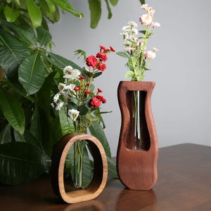 Könnte beinhalten: Zwei Holzvasen, eine ovale und eine geschwungene, die jeweils ein Glasröhrchen für Blumen halten. Die Vasen sind warmbraun und mit bunten Kunstblumen gefüllt. Der Hintergrund umfasst grünes Laub.