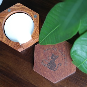 May include: Hexagonal wooden coffee coaster with a cat design and the text "ANINE COFFEE". A white ceramic coffee dripper sits in a wooden holder. The items are on a dark wooden surface.