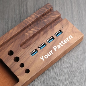 May include: A wooden desk organizer with a brown finish and a built-in USB hub. The organizer has a slot for pens and pencils, a phone stand, and a space for a laptop. The USB hub has four ports. The organizer has the text "Your Pattern" engraved on the side.